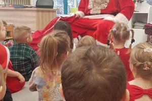 Mikołaj siedzi przy choince, przed nim dzieci